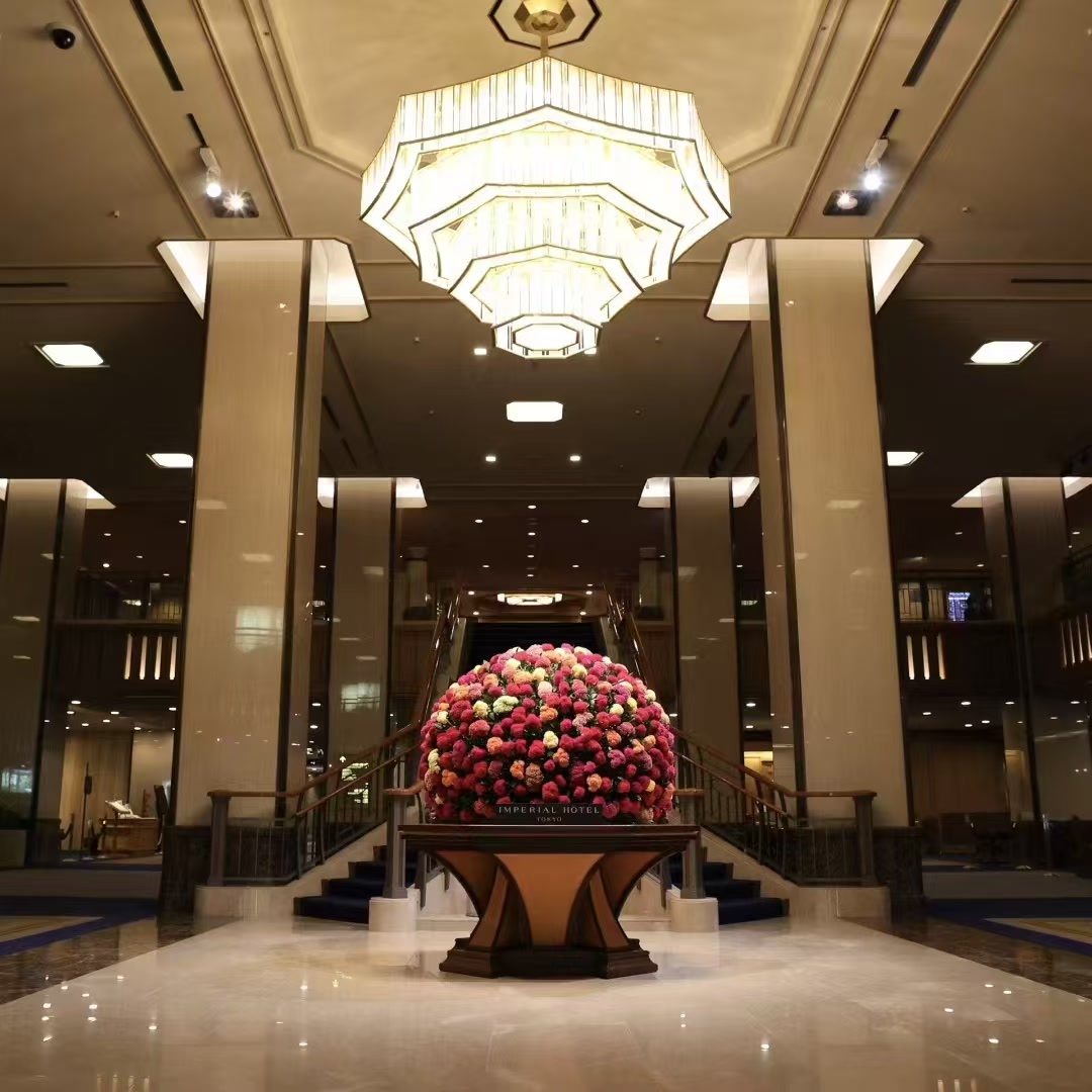 东京帝国饭店正面大堂中央的季节花艺陈设，花艺置于台座上，后方为双侧楼梯与上方灯具